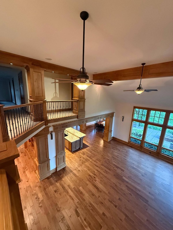 Open living area with clean hardwood floors, wood-trimmed openings, stair railings, ceiling fans, and large windows.