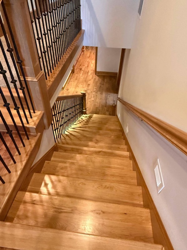 Clean hardwood staircase with polished-looking treads, wood handrails, trim, and black metal balusters connecting the home&rsquo;s levels.