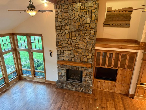 Clean living room area with hardwood floors, stone fireplace, built-in wood cabinetry, and expansive windows.